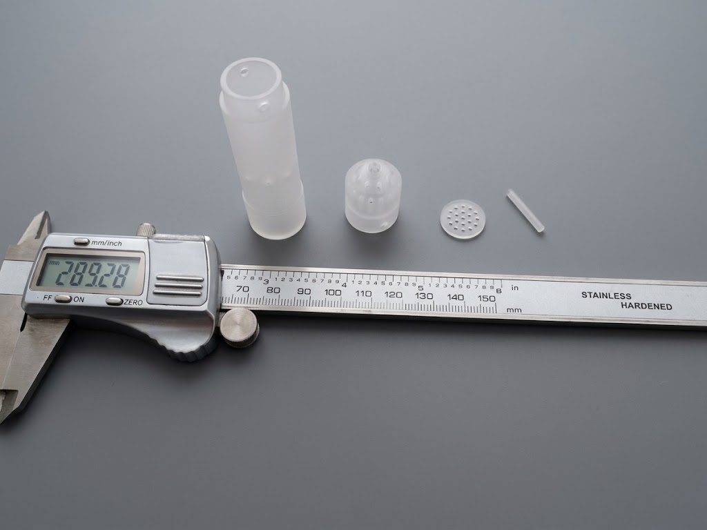 Digital caliper measuring a set of custom-shaped blanks — fluted housing, finned cap, perforated disc, and pin